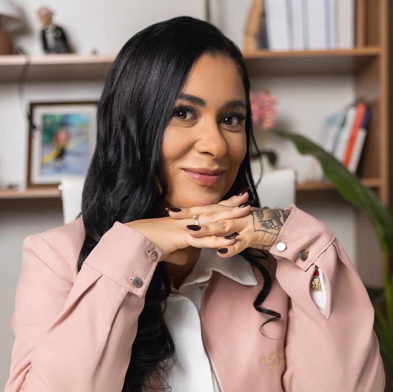 Monique Fahel sorrindo, apoiada na mesa do consultório
